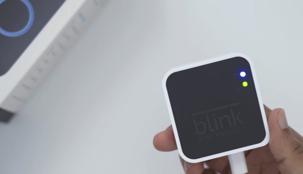 A hand holding a Blink Sync Module 2 with LED indicators lit, indicating connectivity and power